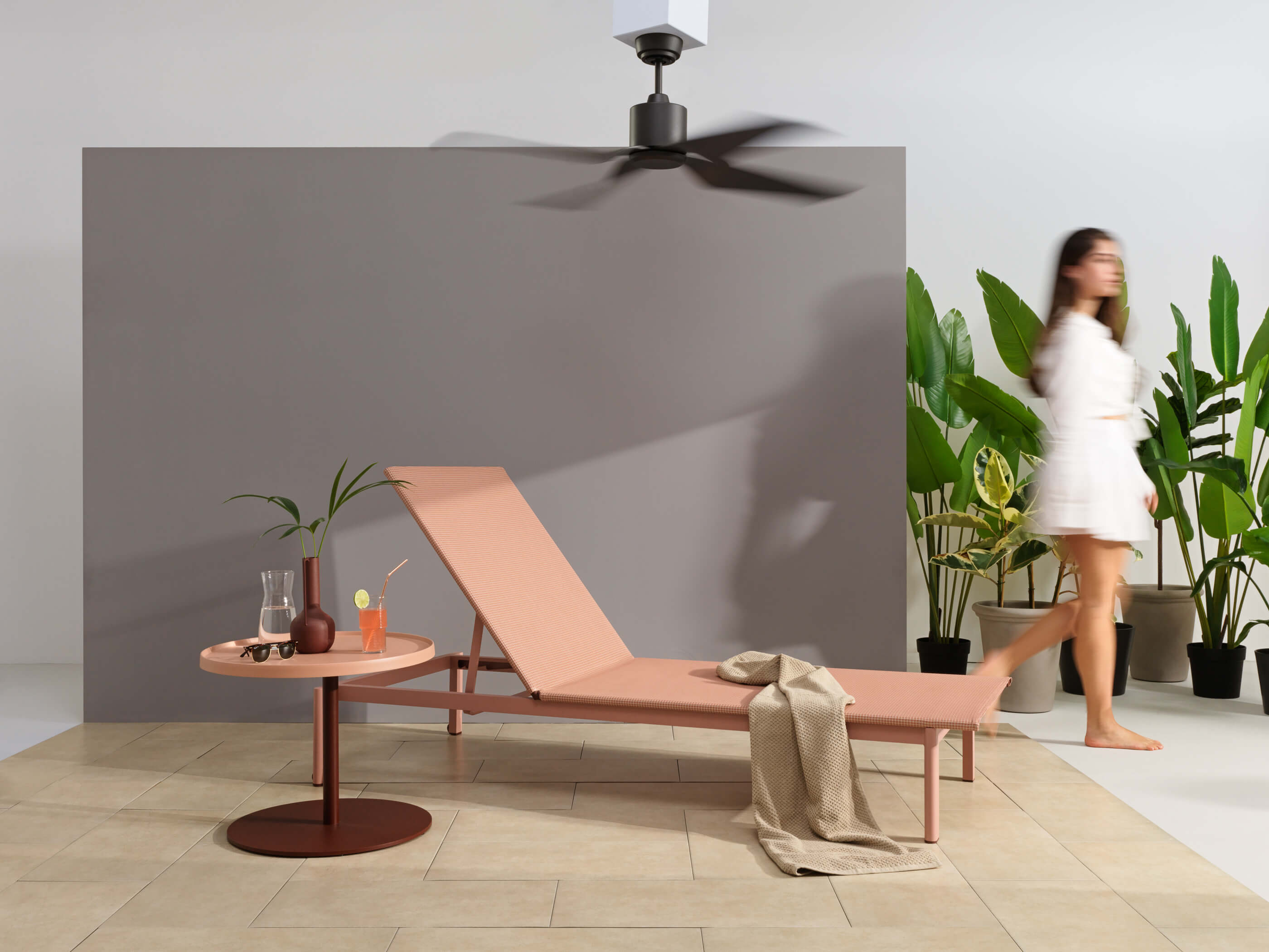 Eine Terasse mit Ventilator, Liege und Pflanzen im Hintergrund, eine Frau läuft vorbei